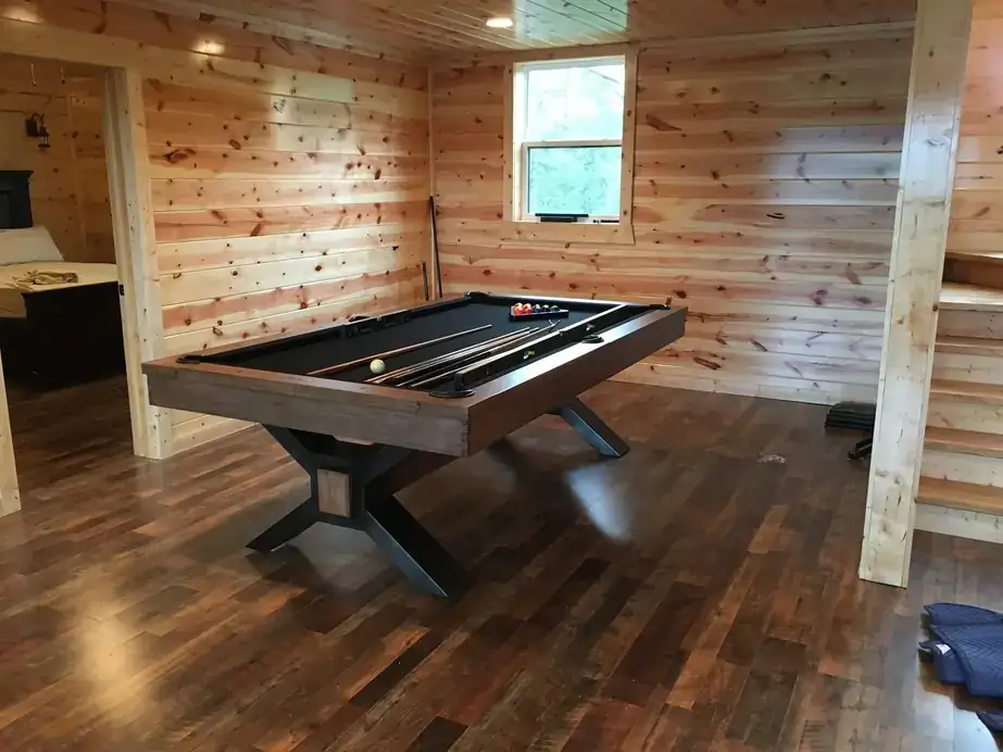 A black pool table with an X-base in a pine-paneled room