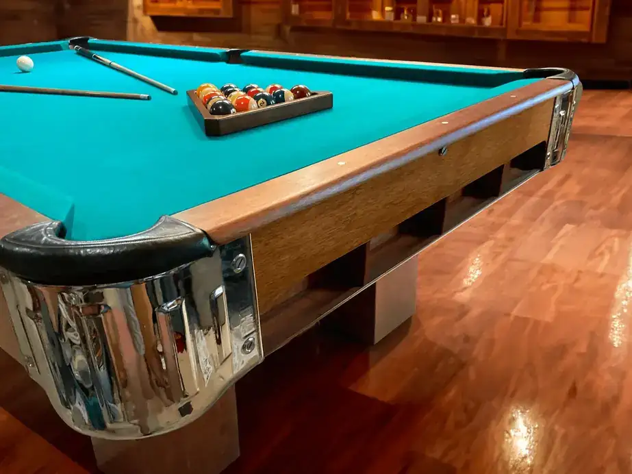 Corner of a teal-cloth pool table with a leather pocket, chrome corner casting and walnut rail