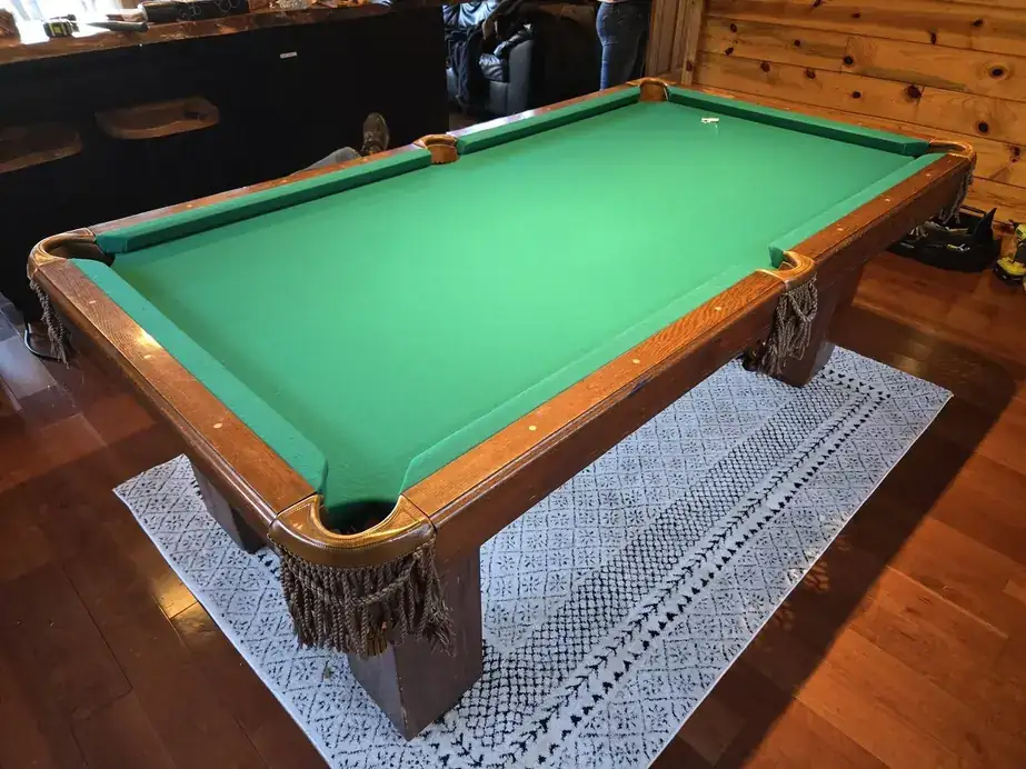 A green-cloth pool table with fringed leather pockets on a patterned rug