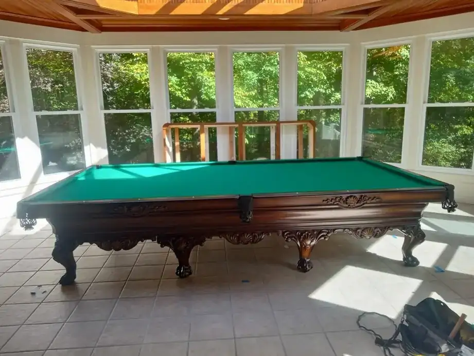 A carved antique pool table with green cloth set up in a bright sunroom